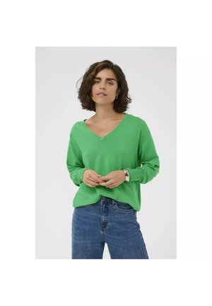 Vrouw met kort krullend haar, gekleed in een groene V-hals trui, blauwe spijkerbroek, creolen en een zilveren armband, staand tegen een witte achtergrond.