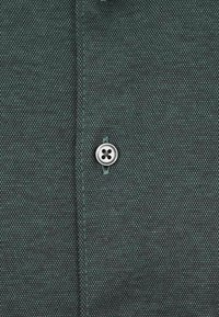 Donkergroene gestoffeerde stof met een subtiel geometrisch ontwerp, met in het midden een zilveren viergaatjesknop.