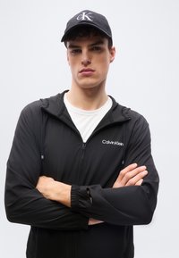 Jeune homme les bras croisés portant une veste zippée noire Calvin Klein, un t-shirt blanc et une casquette noire Calvin Klein sur un fond clair uni.