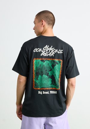 Mann trägt ein schwarzes T-Shirt mit grünem Kaktusmotiv, umrahmt von orangefarbener Stacheldraht und dem Text "CONDITIONS GEAR" sowie "Big Bend, TEXAS" auf dem Rücken.