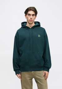 Sweat à capuche turquoise en tissu doux, doté d'une poche kangourou et d'une capuche réglable avec cordon. Un petit patch logo est situé sur la poitrine gauche.