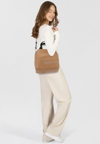 Sac fourre-tout en cuir tan avec poignées noires, ferrures en argent et logo imprimé. Texture lisse et forme carrée, adapté à un usage décontracté.