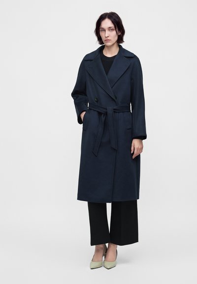 Femme portant un trench coat bleu marine foncé ceinturé, un pantalon noir à jambes larges et des chaussures pointues de couleur claire, debout devant un fond uni.