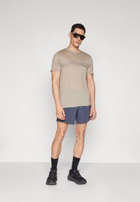Beige t-shirt with a graphic mountain design in multi-colored lines; paired with navy shorts and black sports shoes, worn with sunglasses.