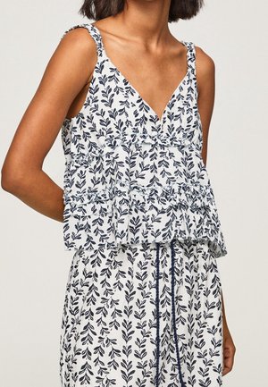 Kvinde iført hvid ærmeløs top og bukser med navyblåt bladmotiv og flæser på toppen.