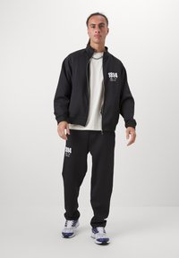 Svart zip-up jakke og matchende joggebukser med hvit '1894'-tekst. Antrekket har en avslappet passform og er laget av glatt, teksturert stoff.