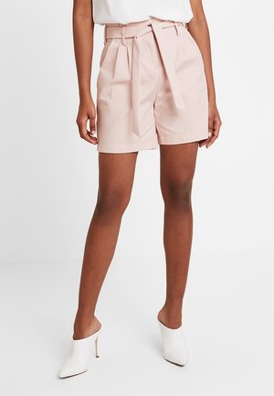 Femme portant un short paperbag taille haute rose clair avec ceinture, associé à des mules blanches à talons hauts et un haut blanc.