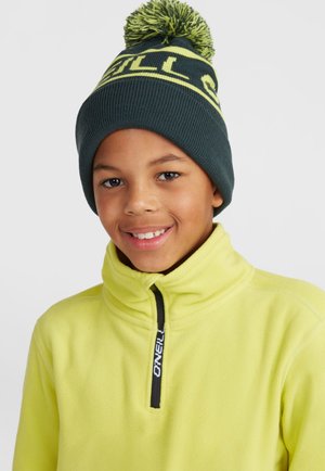Donkergroen gebreide beanie met een gele pompon en opdruk aan de voorkant. Draagt een felgele fleece jas met hoge kraag.