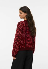 Pull en tricot rouge avec des motifs de texte répétés "LOVE" et "AMOUR", à manches longues et à coupe décontractée. Un pantalon noir est porté en dessous.