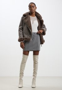 Giacca in pelle foderata in shearling marrone, body a righe bianche, mini gonna grigia con bottoni e stivali alti fino al ginocchio color crema con tacco.