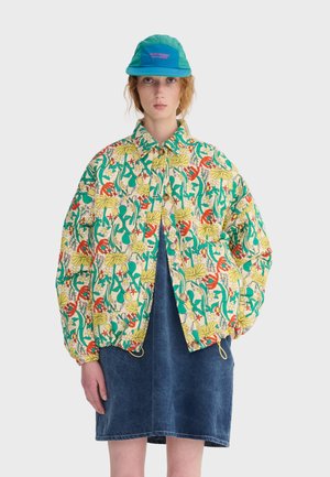 Kvinde iført en lys jakke med blomsterprint, blå denimkjole og teal kasket foran en ensfarvet lys baggrund.