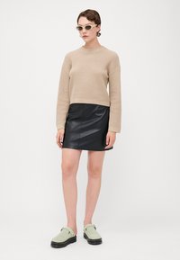 Beige strikkegenser, svart syntetisk skinnskjørt og grønne semskede slip-on-sko. Solbriller fullfører utseendet. Enkelt design med glatte teksturer.