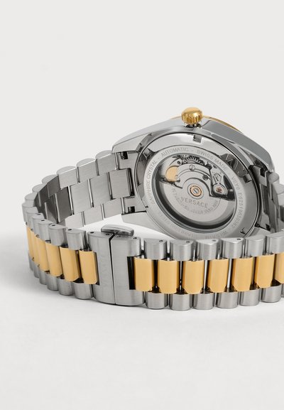 Bracelet de montre en métal argenté et doré avec mouvement automatique visible à travers le fond transparent marqué Versace.
