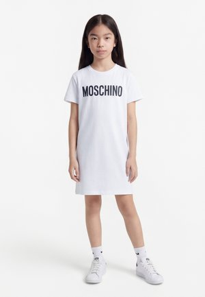Νεαρή κοπέλα στέκεται, φορώντας ένα λευκό φορέμα T-shirt Moschino, λευκές κάλτσες Nike και λευκά αθλητικά παπούτσια, μπροστά σε απλό λευκό φόντο.