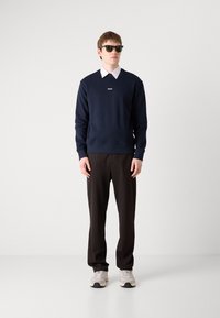 Marineblå genser med hvit logo, kombinert med en hvit skjorte med krage, svarte bukser og grå joggesko. Modellen har på seg svarte solbriller.