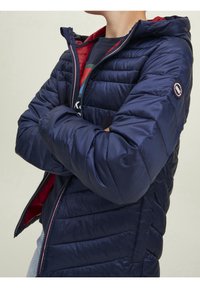 Námořnická péřová bunda s kapucí, která má prošívaný design, červené podšívání a zipové uzavírání. Doplněná logem na rukávu.