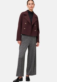 Donna che indossa un cappotto corto doppiopetto di un intenso bordeaux scuro, un dolcevita nero, pantaloni grigi a gamba larga e mocassini neri su uno sfondo chiaro.