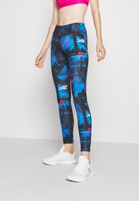 Kvinna i blå och svarta mönstrade leggings, en rosa sport-bh och vita träningssneakers, stående mot en enfärgad bakgrund.