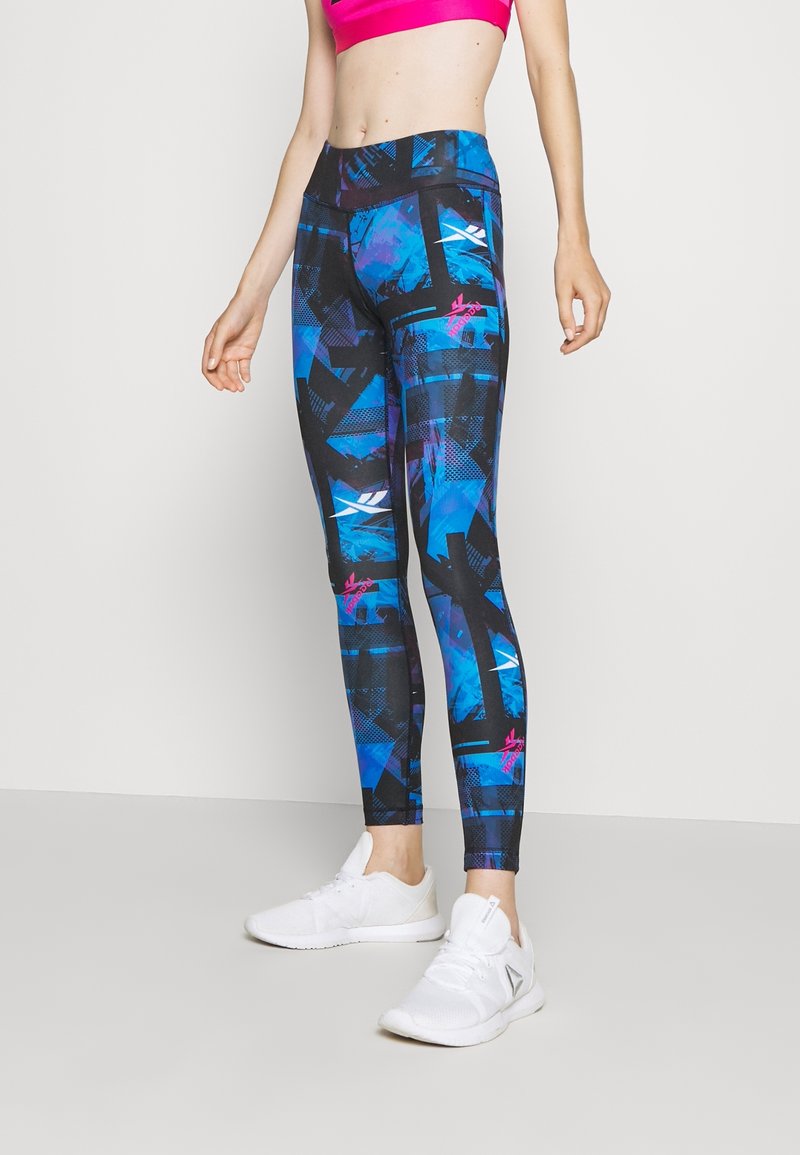 Kvinna i blå och svarta mönstrade leggings, en rosa sport-bh och vita träningssneakers, stående mot en enfärgad bakgrund.