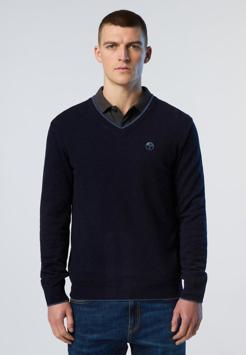 Marineblaues V-Ausschnitt-Pullover aus weichem Strick, mit einem blauen Akzent am Kragen, getragen über einem dunkelgrauen Hemd mit Kragen und kombiniert mit Jeans.