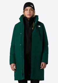 Grøn isoleret jakke med høj krave, lynlås foran og trykknapper. Har et sort fleece indre lag og logo på brystet.