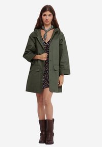 Veste imperméable vert olive avec capuche, fermetures à pression et deux poches avant, portée par-dessus une robe à fleurs, associée à des bottines marron.