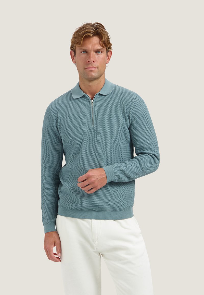 Homme aux cheveux ondulés portant un pull bleu à col zippé manches longues et un pantalon blanc sur un fond clair uni.