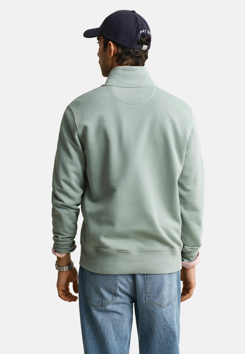 Homme portant un pull vert clair, un jean bleu et une casquette de baseball bleu marine, debout de dos face à l'appareil photo sur un fond blanc.