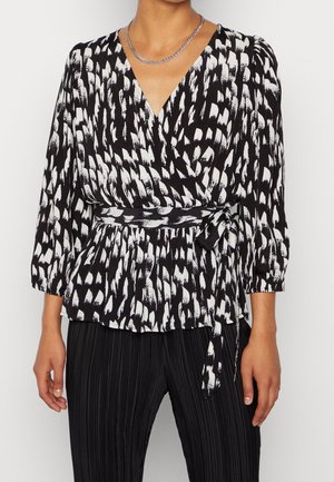 Zwart-wit gebloemde wikkelblouse met driekwart mouwen en een strik in de taille, gecombineerd met zwarte geplooide broek en een zilveren ketting.