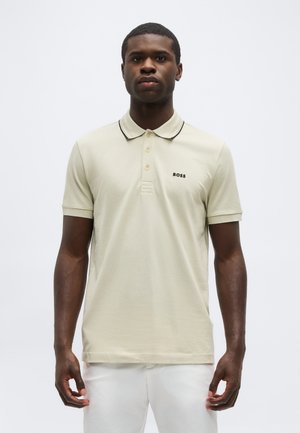 Uomo che indossa una polo beige a maniche corte con colletto bordato di nero e piccolo logo "BOSS" sul petto a sinistra, abbinata a pantaloni bianchi.