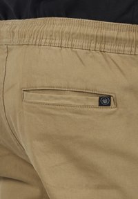 Närbild av beige byxor med elastisk midja och bakficka med bakfickslapp som har en liten svart logotyp.