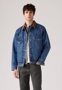 Joven vestido con una chaqueta de mezclilla azul con cuello marrón, camisa a cuadros debajo y jeans gris oscuro, de pie contra un fondo liso.