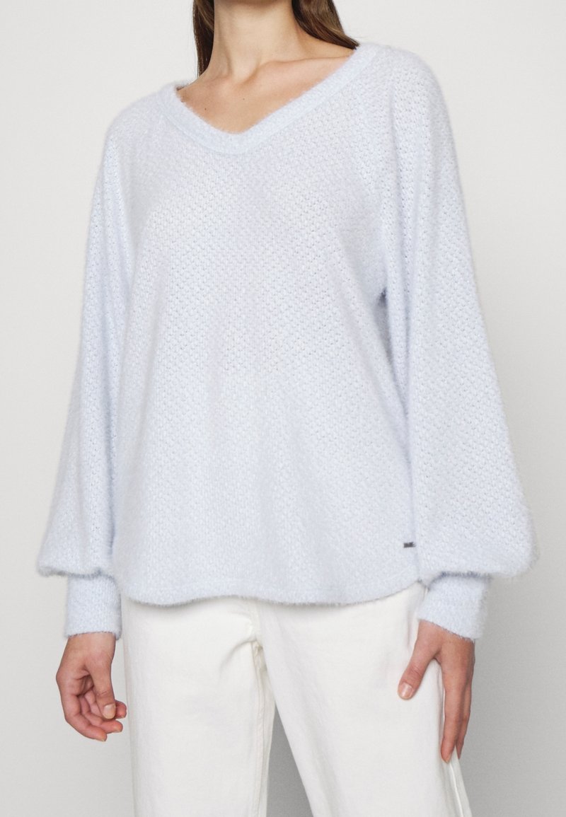 Ljusblå stickad tröja med V-ringning, oversize design och ribbade muddar. Mjuk textur med subtilt mönsterdetalj. Stylad med vita byxor.