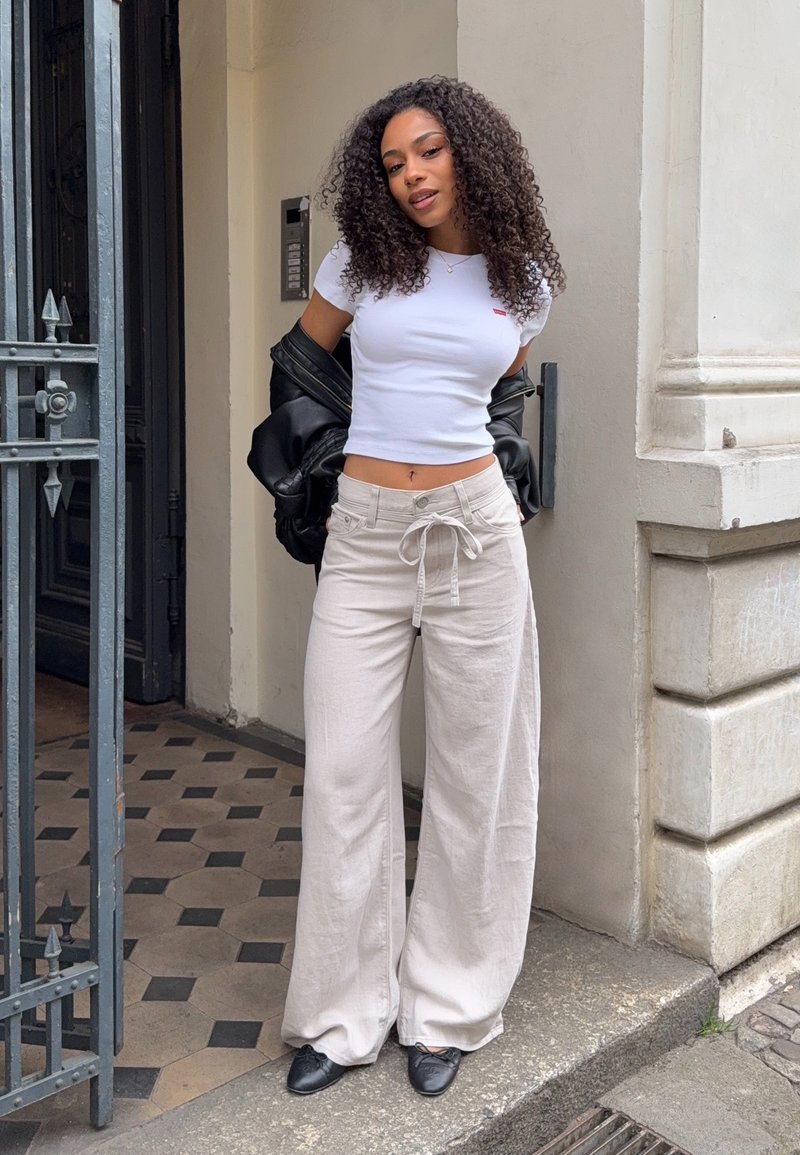 T-shirt bianco corto con logo rosso, pantaloni beige a gamba larga con coulisse, giacca di pelle nera e scarpe nere. Capelli ricci che incorniciano il viso.