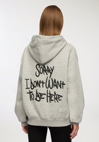 Felpa grigia in cotone con un grande design testuale nero sul retro che recita "MI DISPIACE, NON VOGLIO ESSERE QUI." Presenta un cappuccio con cordino e polsini a costine.