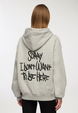 Felpa grigia in cotone con un grande design testuale nero sul retro che recita "MI DISPIACE, NON VOGLIO ESSERE QUI." Presenta un cappuccio con cordino e polsini a costine.