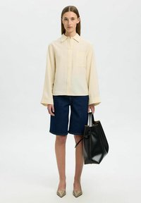 Camisa de botones de manga larga crema, pantalones cortos de mezclilla tipo Bermuda, tacones de punta beige y un bolso tipo cubo negro con un detalle de cinta.