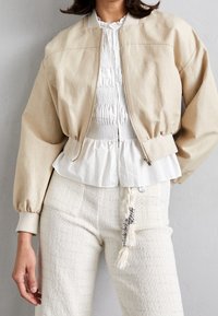 Beige cropped bomberjack met geribbelde manchetten, over een witte plisserok blouse en textuur witte broek met een decoratieve kwastdetail.