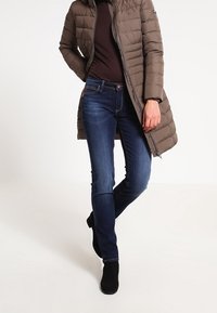 Brązowa puchówka, długa, z zamkiem błyskawicznym, noszona na dopasowanej ciemno-brązowej bluzce. Niebieskie dżinsy z lekkim rozkloszowaniem i czarne botki za kostkę.