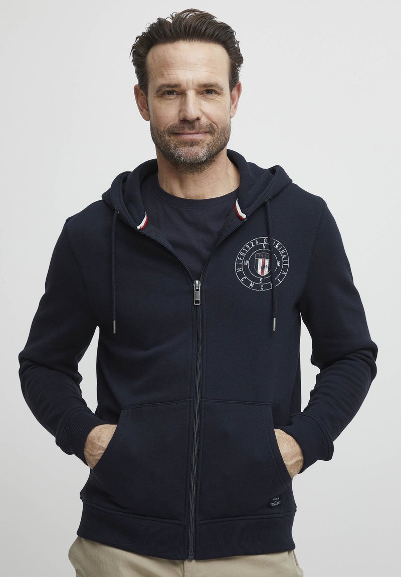 Sweat à capuche zippé bleu marine avec une poche kangourou, une capuche à cordon et un logo sur la poitrine. Comprend un tissu texturé et des poignets côtelés.