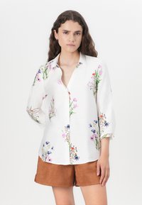 Witte bloes met bloemenprint in roze, paars en groen. Met 3/4 mouwen en een ontspannen pasvorm. Gekoeld met bruine shorts.