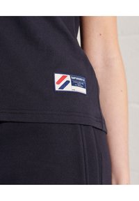 Camiseta de color azul marino con un acabado texturizado, que presenta un parche rectangular de la marca Superdry con acentos en rojo, blanco y azul en el dobladillo.