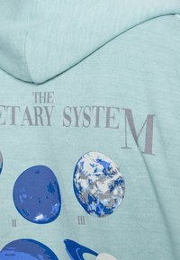 Ljusblå hoodie med ett grafiskt tryck av planeter, med texten "DET PLANETÄRA SYSTEMET M". Mjuk textur, avslappnad design.