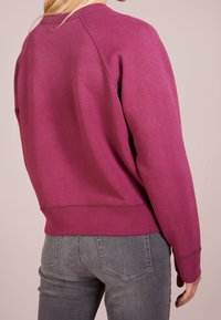 Person som bär en långärmad, magentafärgad sweatshirt tillsammans med grå jeans, sedd bakifrån mot en neutral bakgrund.