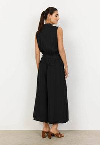 Svart ärmlös jumpsuit med figurnära midja, vida ben och bakfickor. Texturerad tyg, kombinerad med bruna klacksandaler med blockig klack.