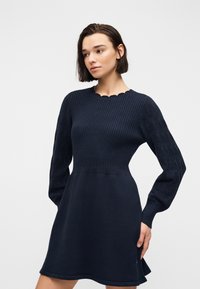 Γυναίκα με κοντά σκούρα μαλλιά, που φοράει μπλε πλεκτό φόρεμα με ριμπ ύφασμα στο μπούστο και φουσκωτά μανίκια με σχέδιο, κοιτάζει ελαφρώς προς τα αριστερά της.