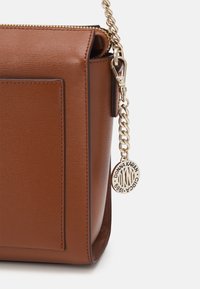 Brun läderhandväska med texturerad yta, en guldkedjestraps och en framträdande varumärkesetikett. Har en dragkedjestängning och en platt framficka.