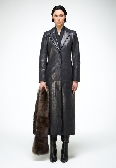 Femme debout portant un long manteau en cuir noir et des bottes, tenant une longue écharpe en fourrure brun foncé sur un fond blanc uni.