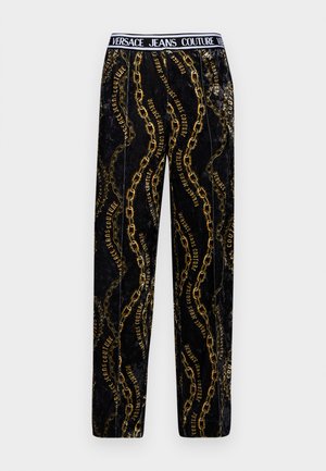 Czarne aksamitne spodnie z złotymi łańcuchami i wzorami tekstowymi, z czarnym pasem oznaczonym "Versace Jeans Couture". Prosty krój nogawki.