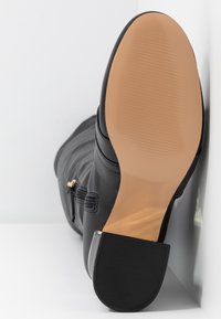 Botte en cuir noir reposant sur le côté montrant une semelle lisse marron clair et un talon noir bas avec un détail de fermeture éclair sur le côté.